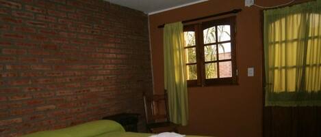 A room with a brick wall, a bed with a green bedspread, a window with yellow curtains, and a wooden chair.