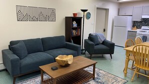 Un salon comprenant un canapé, un fauteuil, une table basse, une bibliothèque et un coin cuisine avec des placards et des appareils électroménagers.
