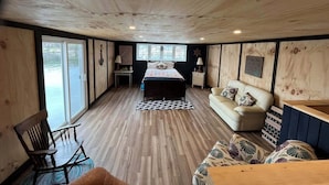 Interior de una cabaña con techo de madera, una cama con colcha estampada, un sofá con cojines decorativos, una mesa de madera y vistas al exterior a través de una puerta corrediza.
