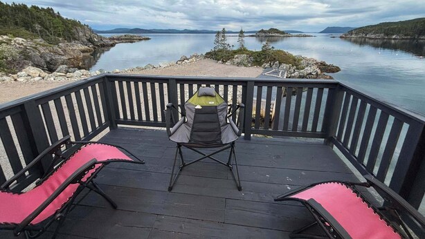 Una terraza con dos sillas y una mesa, con vistas a un lago tranquilo con islas lejanas y un cielo nublado.