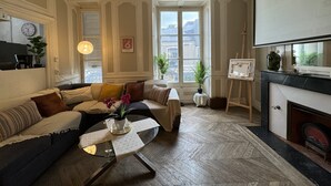 A living room with a sofa, coffee table, and a fireplace. There is a window with a view of buildings outside. - Villa in Le Mans Near Plantagenet City (Le Mans)