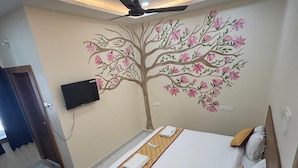 Un dormitorio con cama, ventilador de techo, televisor montado en la pared y un mural decorativo con motivos de árboles.