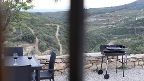 Ein Tisch mit zwei Weingläsern, ein Korbstuhl und ein Grill an einer Steinmauer mit Blick auf eine hügelige Landschaft.