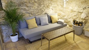 Una sala de estar acogedora con una pared de piedra, un sofá gris con cojines, una planta en maceta, un banco de fibra natural y un cesto de leña.