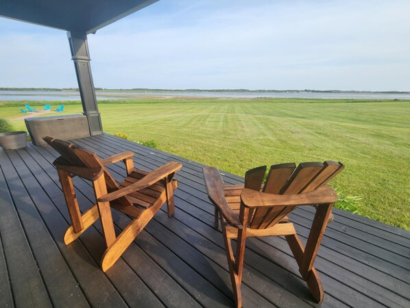 Una terraza de madera con dos sillas Adirondack y una mesa, con vista a un campo cubierto de césped y un cuerpo de agua.