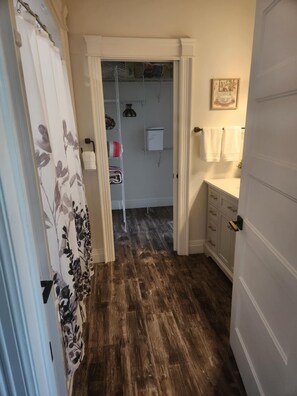 Un pasillo estrecho con piso de madera que conduce a un baño con lavamanos y toallero blancos.