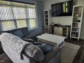 Una sala de estar con un sofá gris, una mesa de centro blanca, un televisor instalado en la pared y estanterías llenas de libros y objetos decorativos.