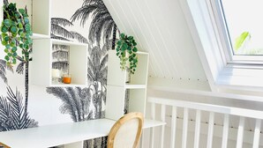Quarto com escrivaninha branca, cadeira, estante e janela com vista para o verde.