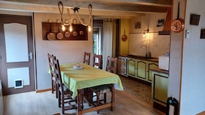 A dining area with a wooden table and chairs, a kitchen with a sink and cabinets, and a chandelier with exposed bulbs. - Gîte Lisière d'Argonne (Passavant-en-Argonne)