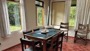 A dining area with a wooden table and chairs, a window with curtains, and a view of greenery outside.