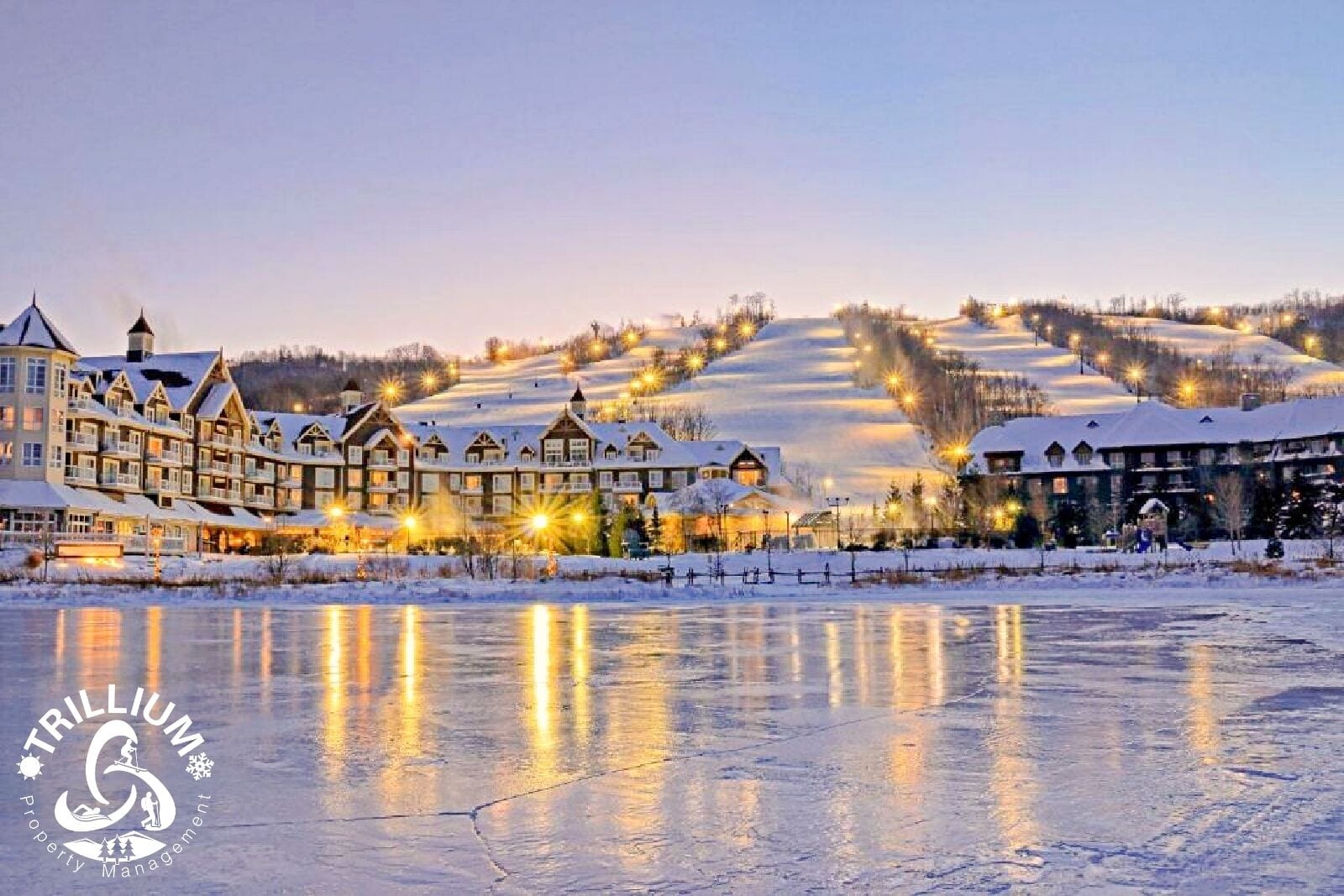 A resort with multiple buildings, a frozen lake in the foreground, and snow-covered hills in the background.