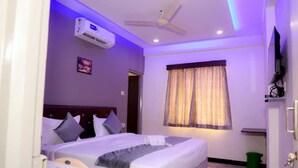 A hotel room with a bed, a ceiling fan, an air conditioning unit, a window with curtains, and a small desk with a chair.