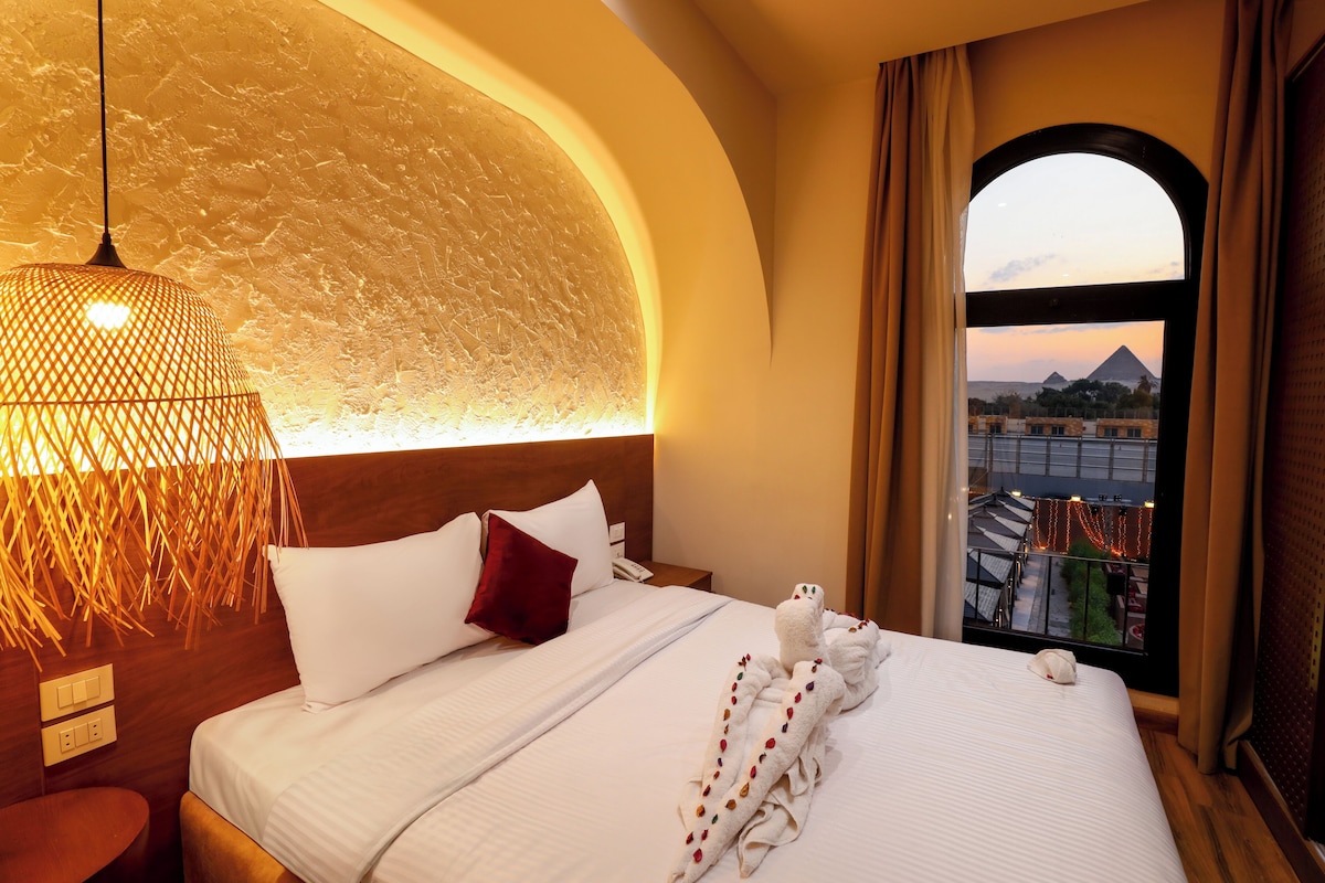 A hotel room with a bed, a nightstand, a lamp, and a view of a pyramid.