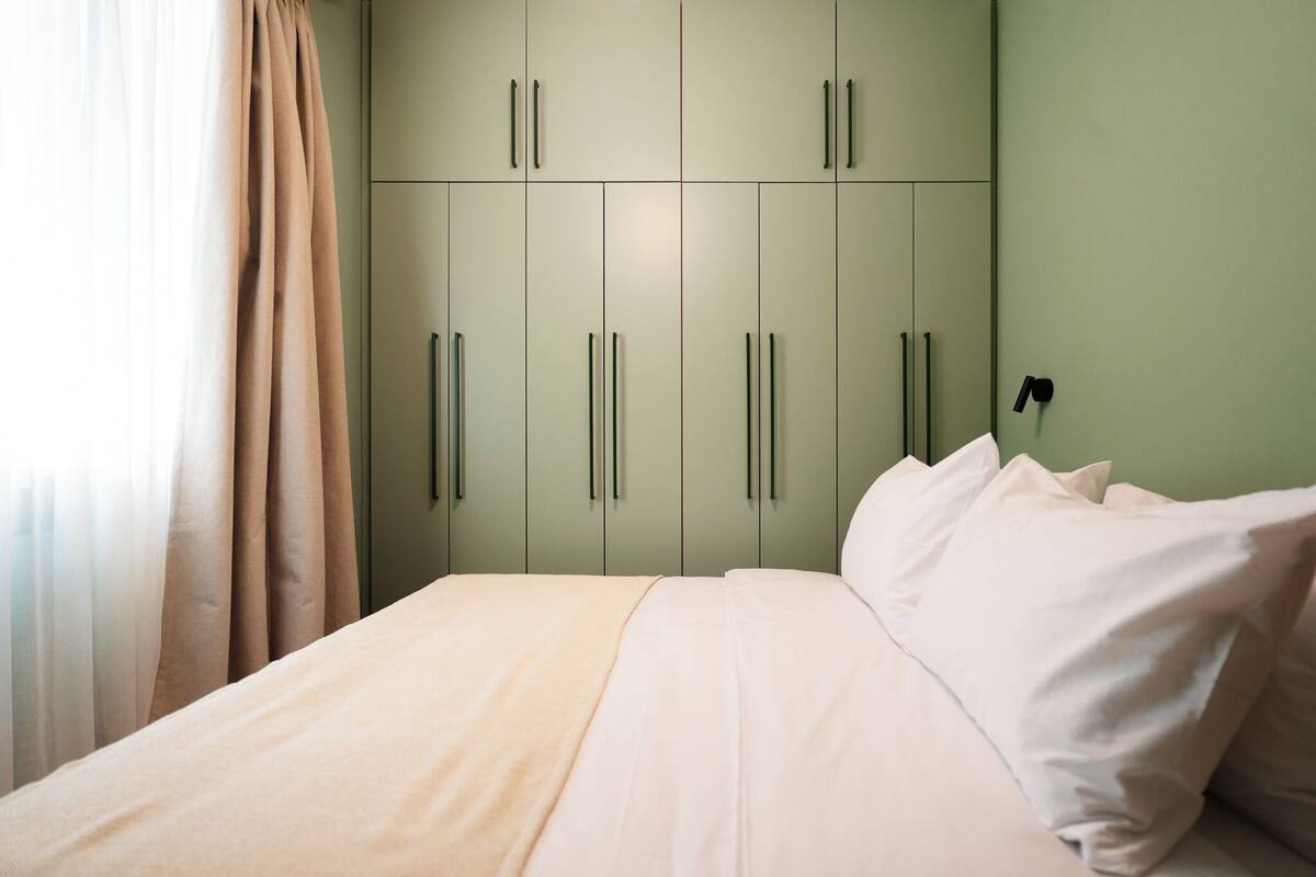 A bedroom with a bed, beige curtains, and a green wardrobe with handles.