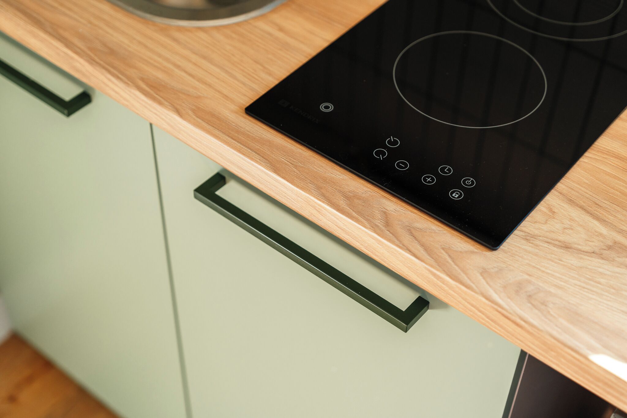 A kitchen with a black induction cooktop, a wooden countertop, and green cabinets with black handles.