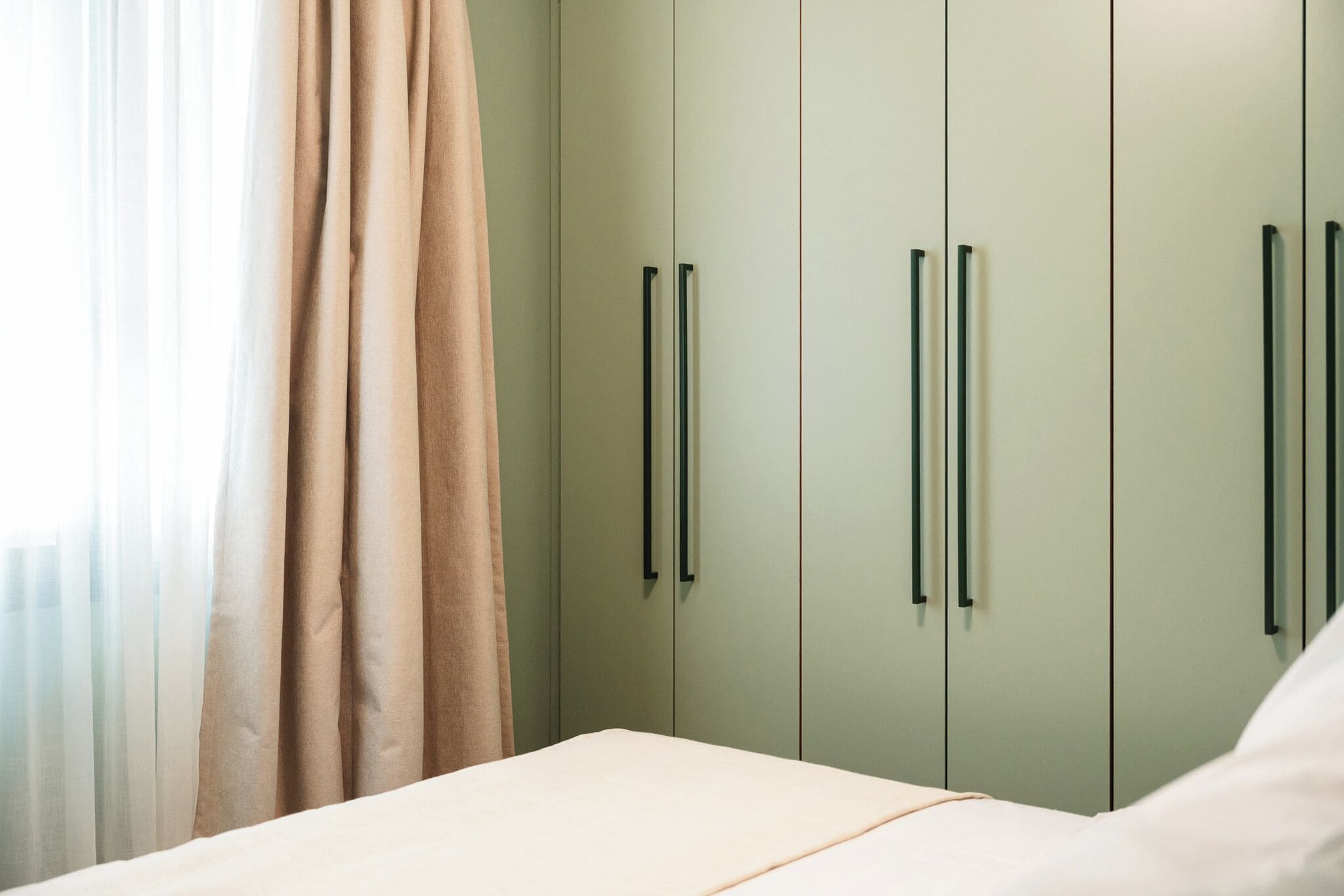 A bedroom with a bed, light green wardrobe with black handles, and sheer curtains.