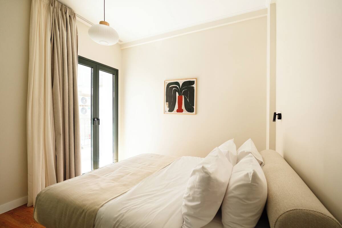 A bedroom with a bed, a window with curtains, a wall-mounted artwork, and a door.