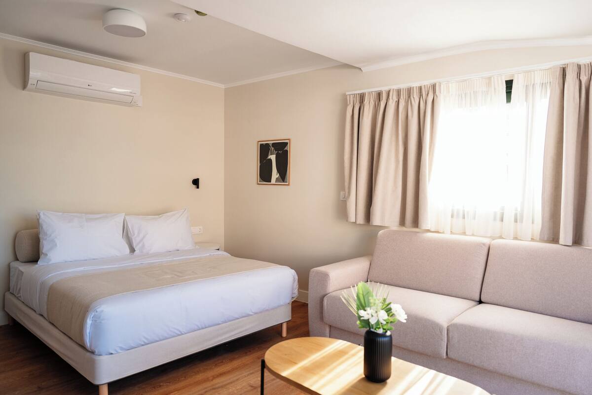 A hotel room with a bed, a sofa, a small table with a vase, an air conditioner, and a window with curtains.