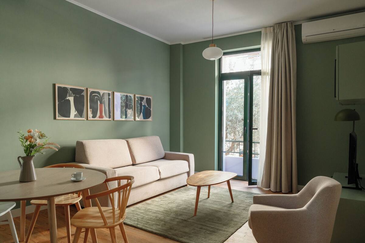 A living room with a sofa, coffee table, and chairs. There are framed pictures on the wall and a window with curtains.