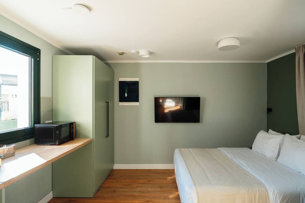 A bedroom with a bed, a TV mounted on the wall, a microwave on a shelf, and a window with blinds.
