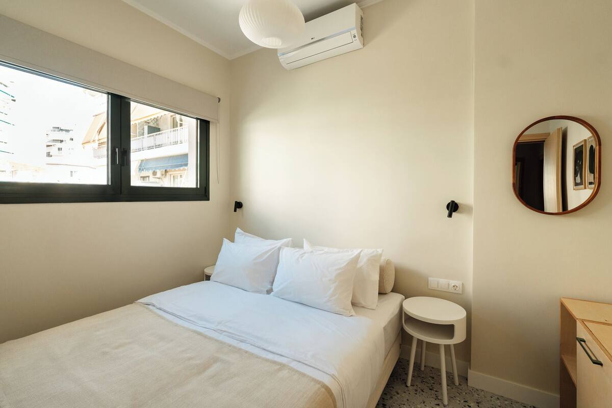 A bedroom with a bed, a small bedside table, a round mirror, and a window with a view of buildings.