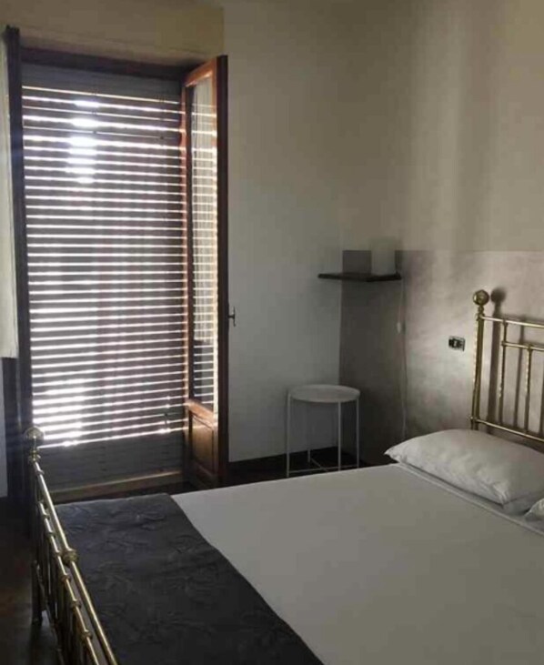 A bedroom with a bed, a small table, a shelf, and a door with blinds. - “Residenza il Castello” 2 BIS (Castell'Arquato)