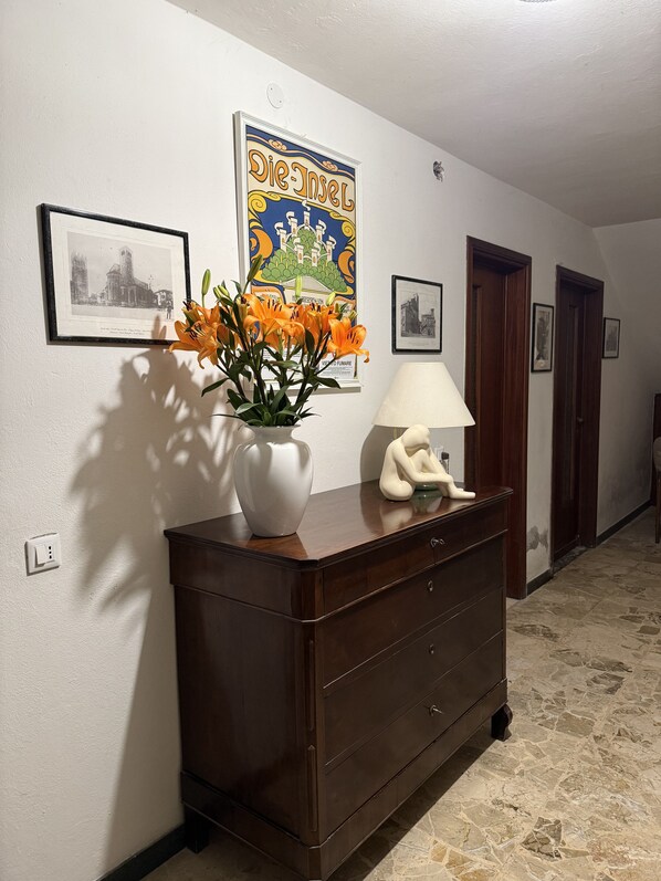 A hallway with a dark wooden dresser, a vase with orange flowers, a framed poster, a lamp, and a statue.