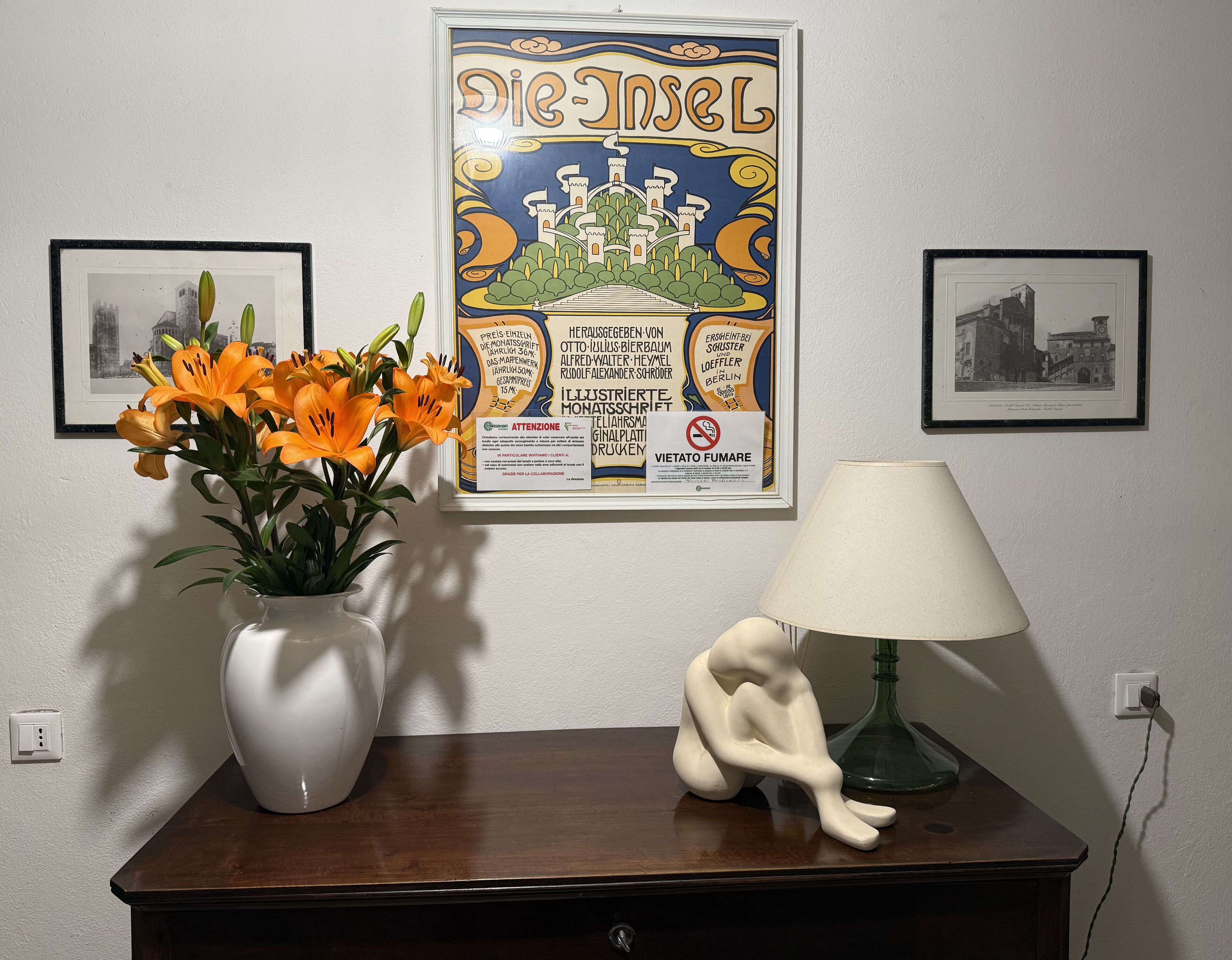 Um cartaz emoldurado com texto e gráficos, um vaso com flores laranjas, uma luminária branca e uma pequena estátua sobre uma mesa de madeira.