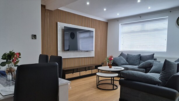 A modern living room with a flat-screen TV mounted on the wall, a grey sofa, a round coffee table, and a dining area with a white table and chairs. - Grand Apartments Stretford (Stretford)