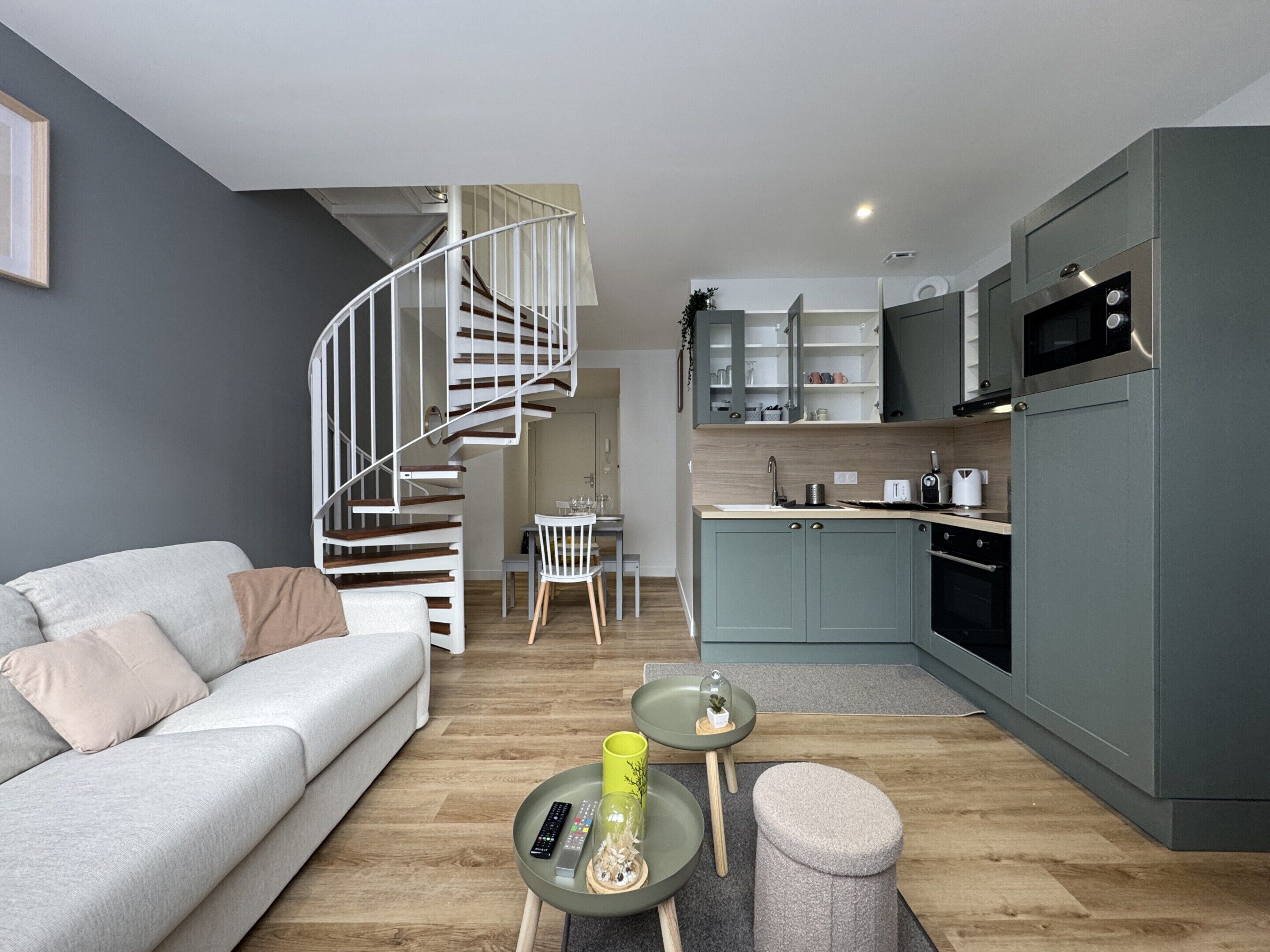Una sala de estar moderna con un sofá gris, una pequeña mesa redonda con un control remoto y un florero de vidrio. Una escalera en espiral conduce a un comedor con una silla blanca y una mesa pequeña. La cocina tiene armarios grises, un microondas y un fregadero.