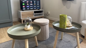 A modern living room with a TV, two small tables, a cushioned stool, and a decorative item on one table. - Duplex in Le Mans Near 24 Hours Circuit (Le Mans)