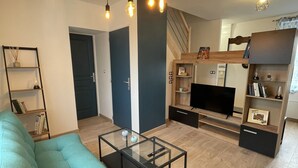 Una sala de estar moderna con un sofá color turquesa, una mesa de centro de vidrio y un mueble de televisión con televisor de pantalla plana. Hay estanterías de madera y una escalera que conduce a un nivel superior.