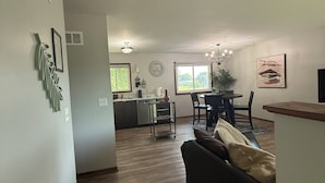 A spacious living area with a dark-colored sofa, a dining table with chairs, a kitchen area with a window, and a chandelier.
