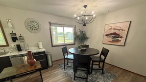 A dining area with a round table, two chairs, a chandelier, a potted plant, a clock, and a window with a view of the outdoors.