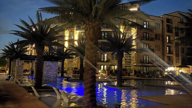 Zona de piscina nocturna con palmeras y un edificio con ventanas iluminadas.