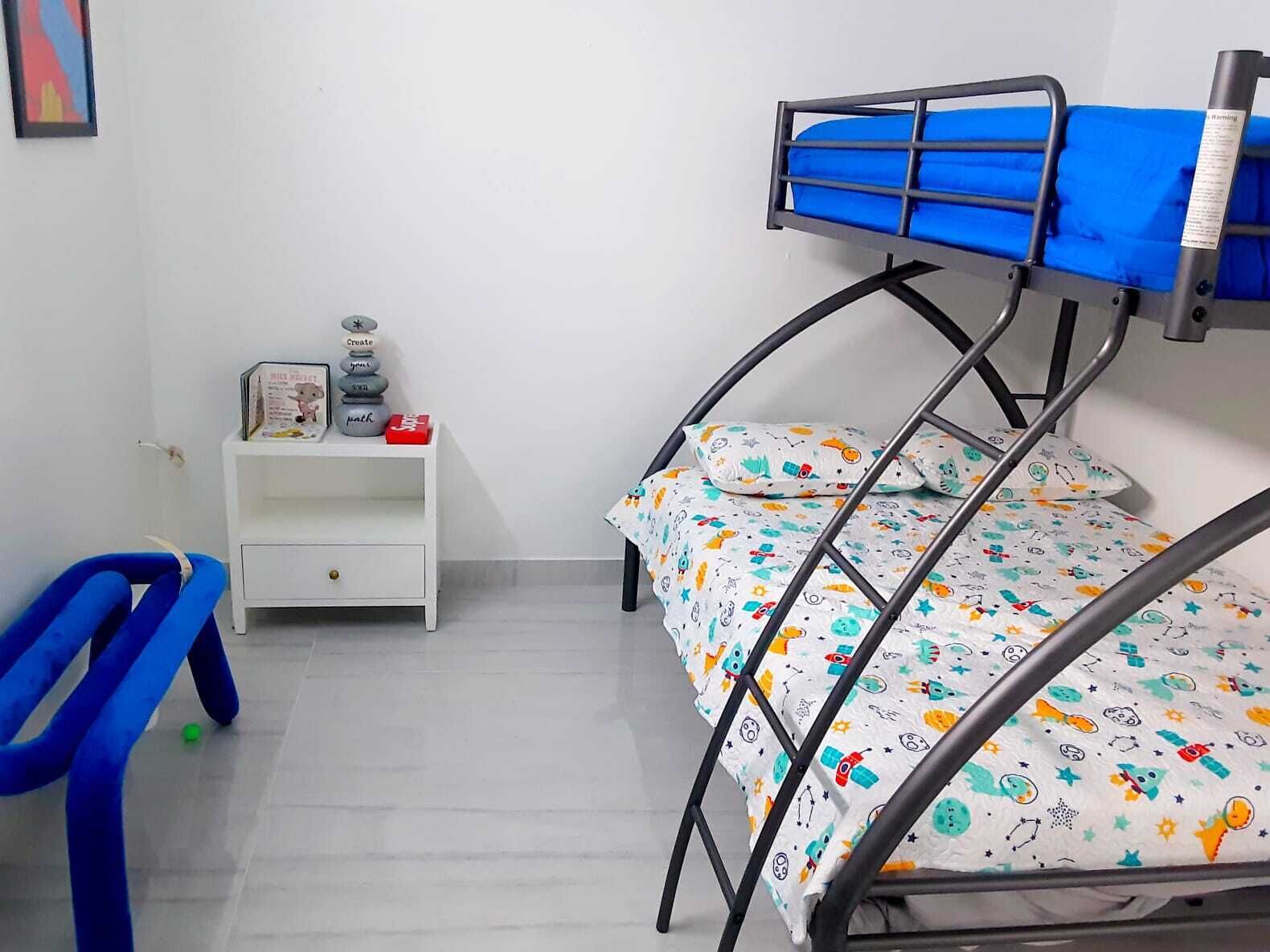 Un lit superposé pour enfant avec une couverture colorée, une table de chevet blanche avec une petite lampe et une chaise bleue.
