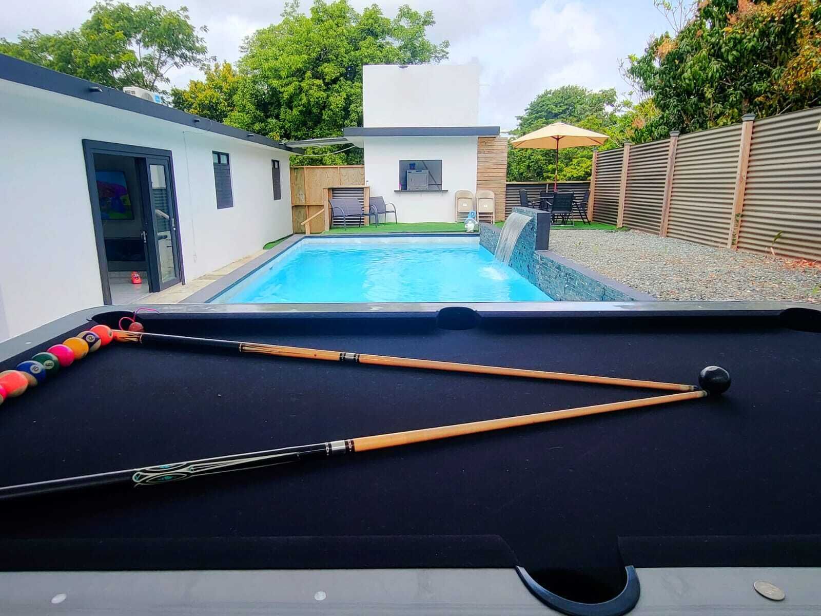 Une table de billard avec des queues et des billes, une maison moderne avec une piscine, et un espace jardin agrémenté d’une clôture en bois et de mobilier d’extérieur.