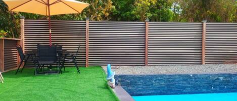 A backyard with artificial grass, a pool, a table with chairs, and a wooden fence.