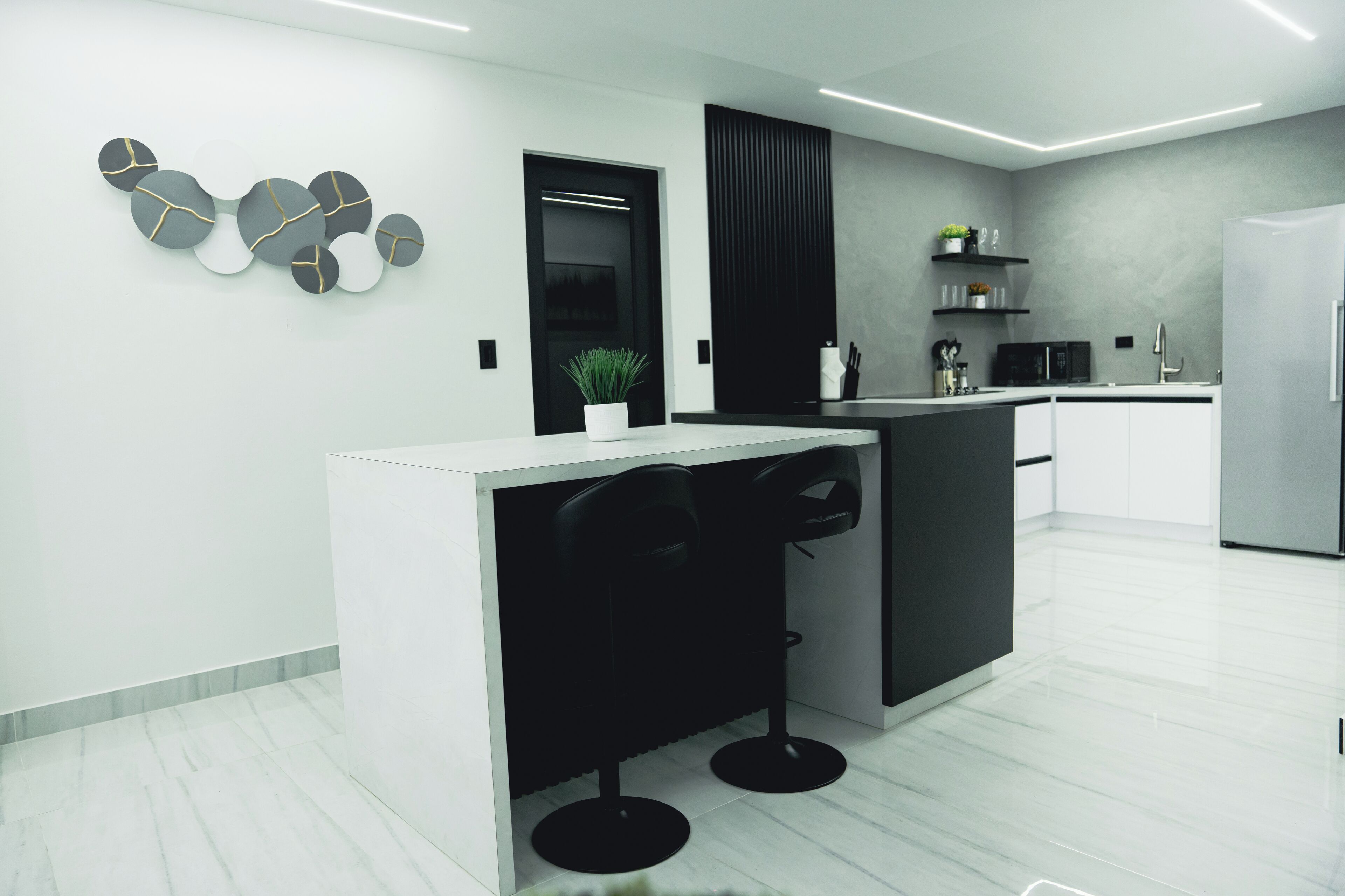 Une cuisine moderne avec un plan de travail blanc, des tabourets de bar noirs, une porte noire et un mur gris agrémenté d’une plante.