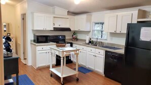 A kitchen with white cabinets, a black refrigerator, a microwave, a stove, and a cart with a basket. - “Our Lakeside Oasis” is a Cozy 2-bedroom house in charming Okeechobee. (Okeechobee)