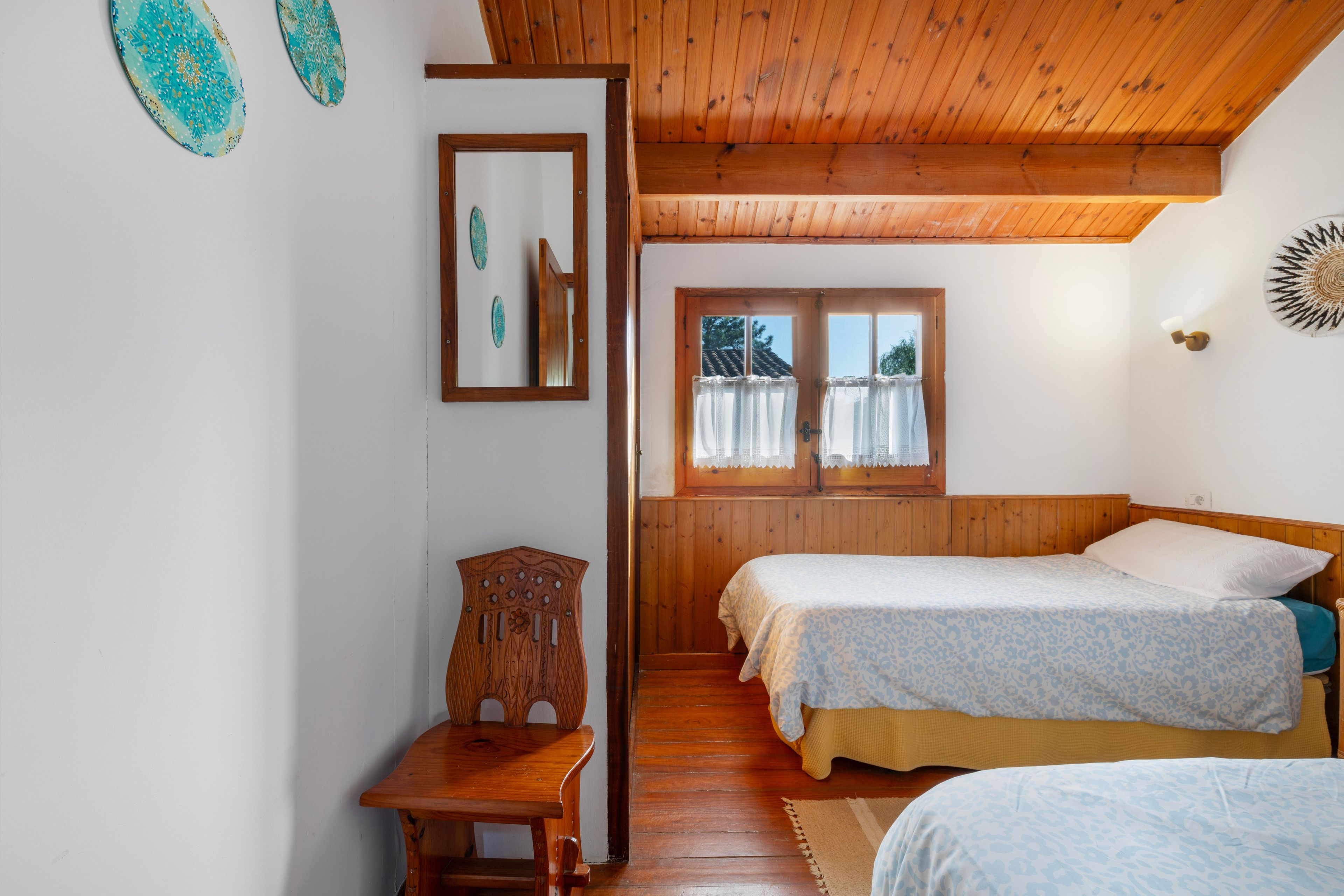 A room with a wooden ceiling, two beds with patterned bedding, a wooden chair, a small table, and a window with curtains.