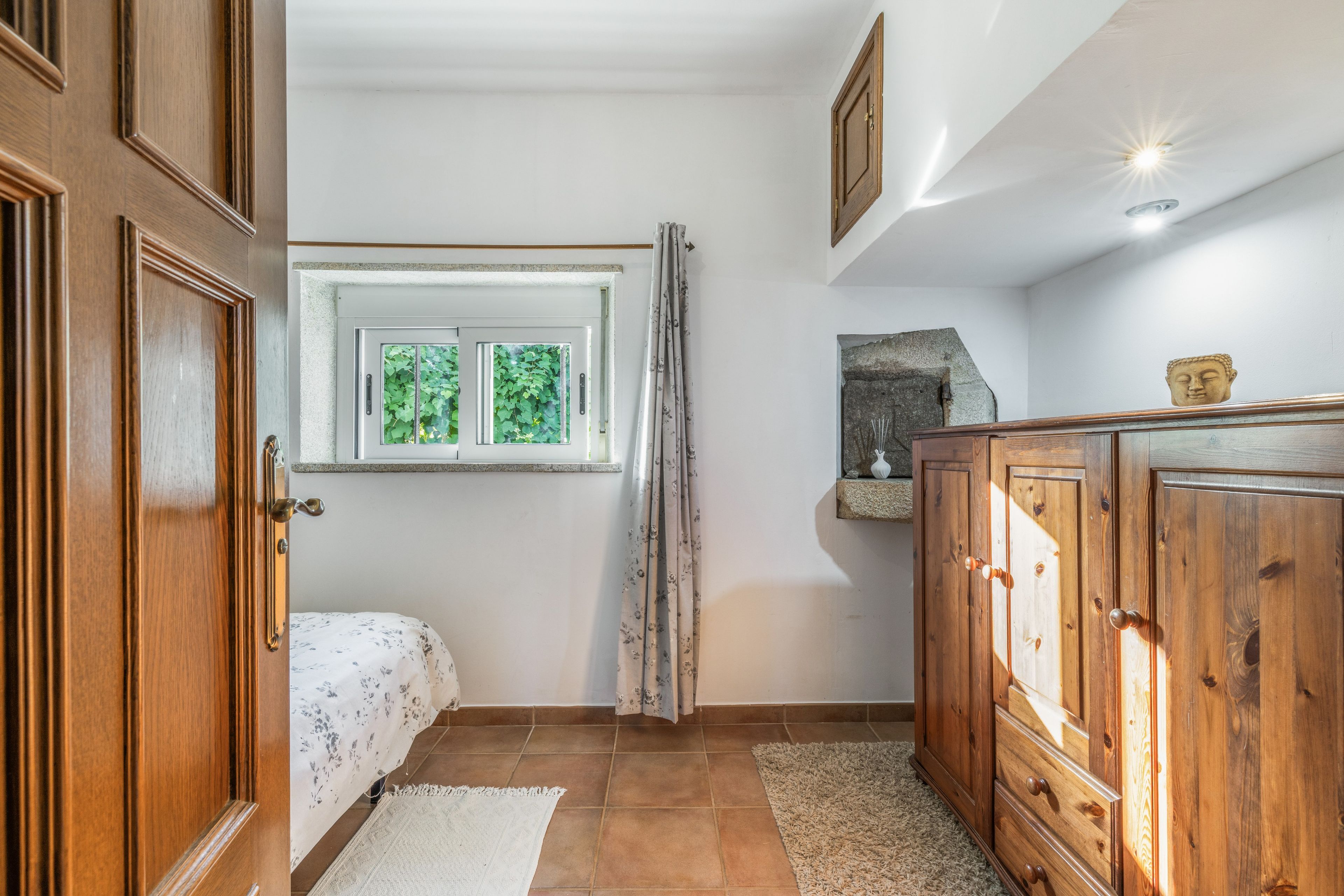Een slaapkamer met een bed, een houten garderobe, een raam met gordijnen, een stenen beeldhouwwerk en een betegelde vloer.