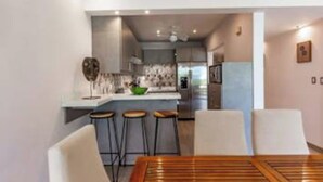 A modern kitchen with a central island, bar stools, and a dining area with a wooden table and chairs. - Elegant Condo with Brand-New Fixtures, Shaded Tropical Terrace, and Secluded Beach Access (Playa Flamingo)
