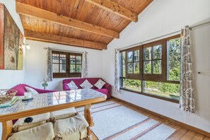 A cozy room with a wooden ceiling, a sofa with cushions, a table with a patterned tablecloth, and a large window with curtains.