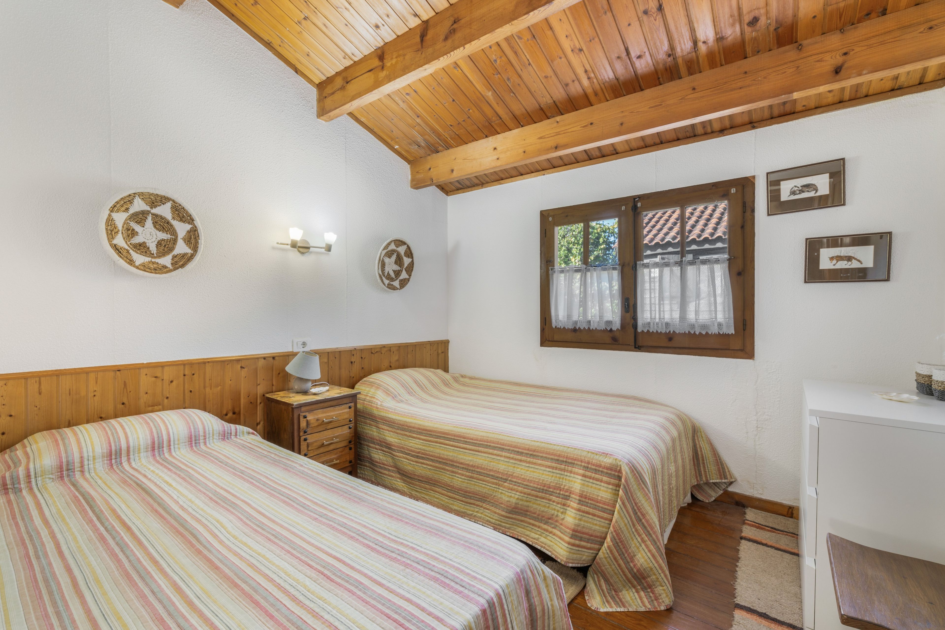 A room with two beds, wooden ceiling beams, a small desk, and a window with curtains.