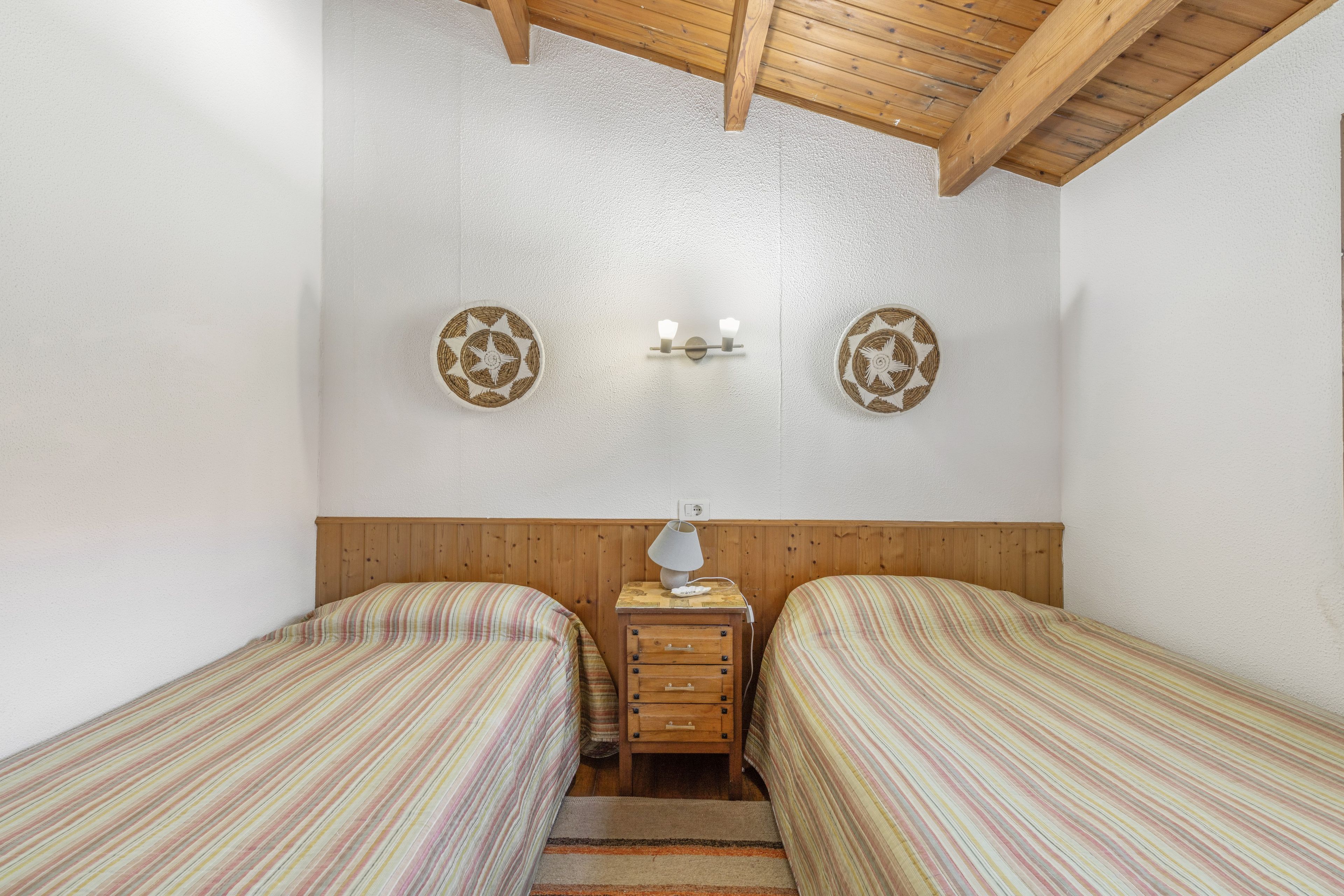 Habitación con dos camas individuales, cabecera de madera, mesita de noche con lámpara y dos luces fijadas a la pared.