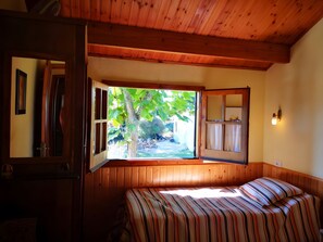 Quarto com cama, janela com vista para vegetação, espelho e luminária na parede.