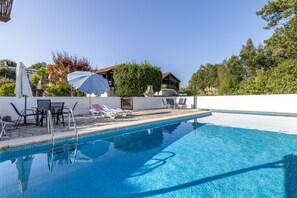 A swimming pool with lounge chairs and umbrellas, surrounded by a white wall and trees.