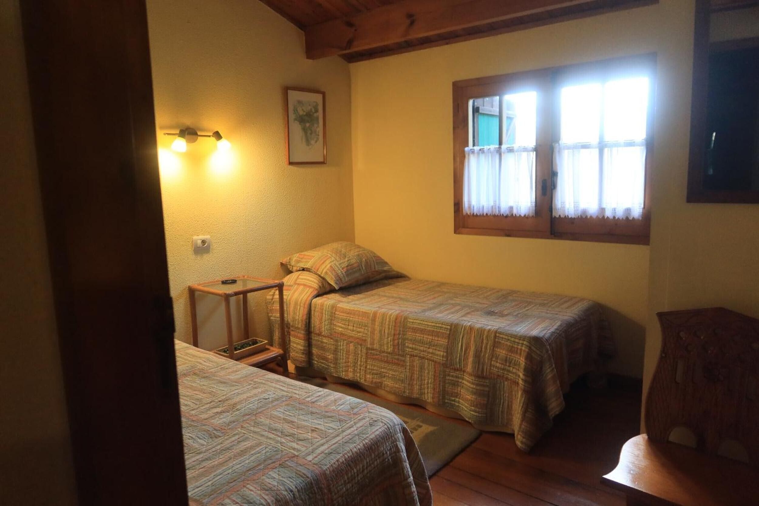 Una habitación con dos camas, un banco de madera, una ventana con cortinas y una lámpara fijada a la pared.