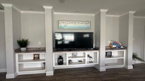 A living room with a flat-screen TV mounted on a white entertainment center, flanked by white shelving units with decorative items and plants.